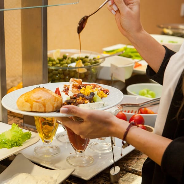 A person drizzles sauce over a breakfast plate with a pastry, cheese, corn, and fruit.